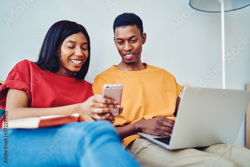 Smiling african american woman showing funny video on smartphone resting together at bedroom in apartment, happy dark skinned young marriage checking social networks on mobile phone enjoy leisure.