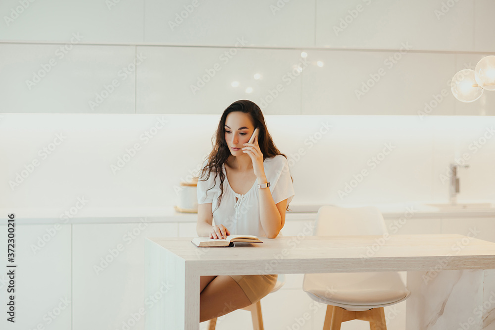 Serious beautiful woman speaks on phone in minimalistic light kitchen Serious beautiful woman speaks on phone in minimalistic light kitchen