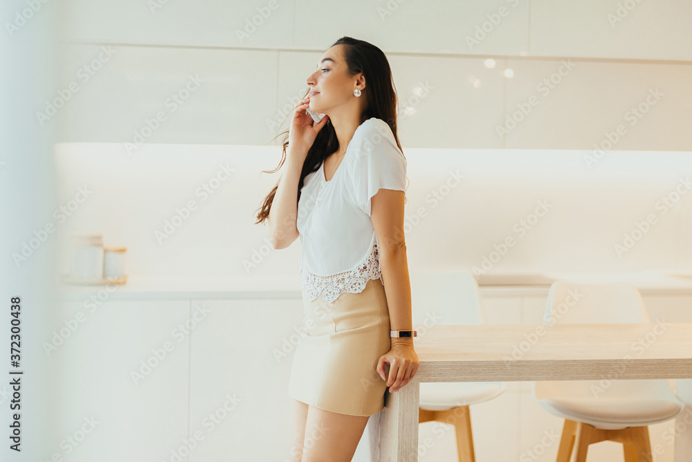 Serious young beautiful woman speaks on phone in minimalistic light kitchen Serious young beautiful woman speaks on phone in minimalistic light kitchen