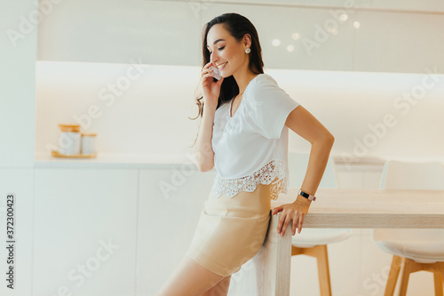 Happy young beautiful woman speaks on phone in minimalistic light kitchen