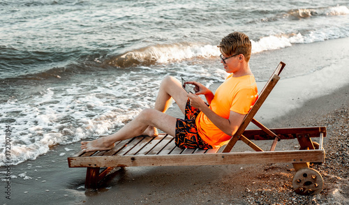 A guy in a T-shirt and shorts holds tablet, sitting on a wooden deck chair by the sea photo