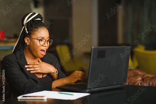 Young girl in glasses amazingly looking in laptop at cafe. African American girl sitting in restaurant with laptop and cup on table. Portrait of surprised lady with dark curly hair in earphones