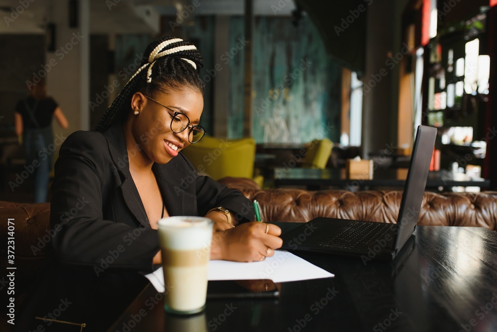 Student girl browsing Internet, using free wi-fi at cafe. African freelancer thinking on ideas for her blog, using laptop at co-working space, resting hand on wooden table, looking with inspired smile