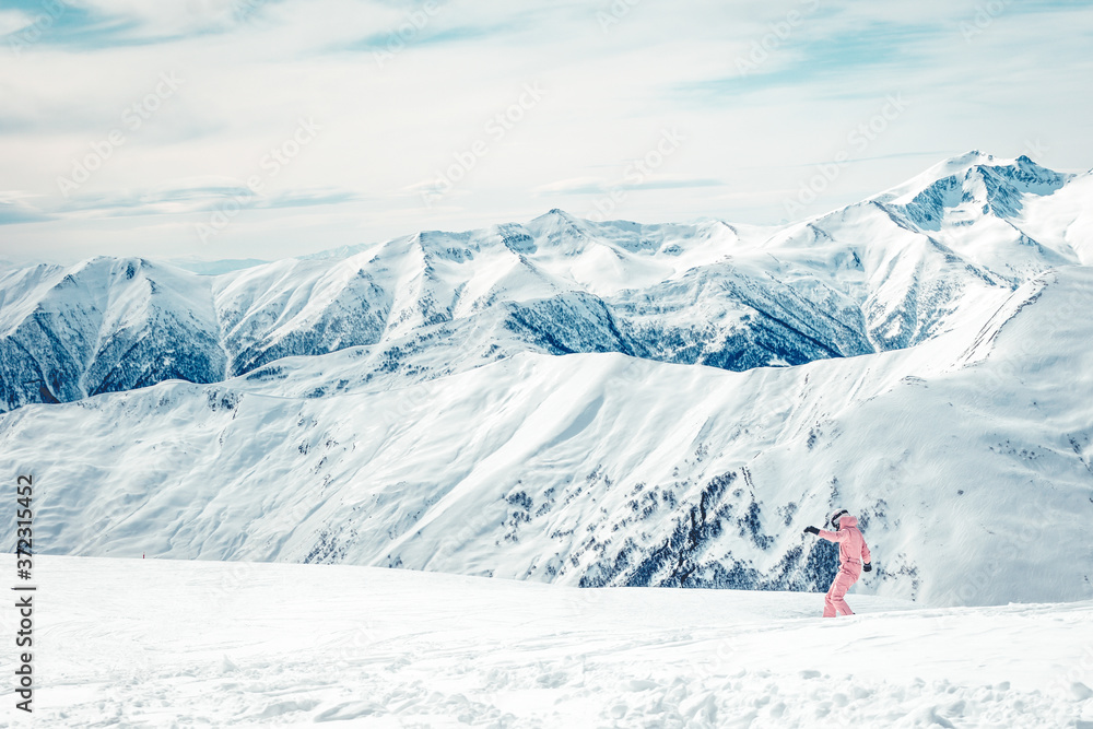 Girl in pink combinezon rides down the hill on snowboard with mountains view in the background.  Skiing holidays in KAzbegi mountains.
