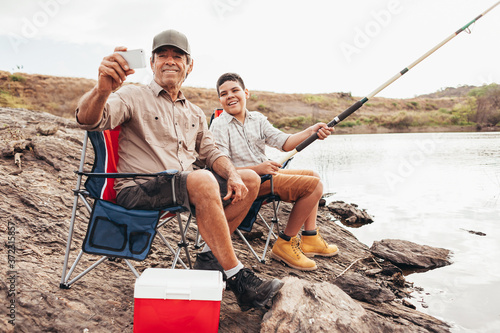 Latin grandfather and grandson enjoying day together fishing on the lake. Taking selfie on smartphone.