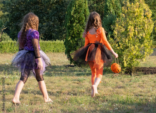 two witch girls go barefoot across the lawn with Jack-o-lanterns for candy. view from the back.