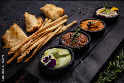 Appetizers sauces with toasted bread and baked sticks are served on wooden stands. On dark concrete 