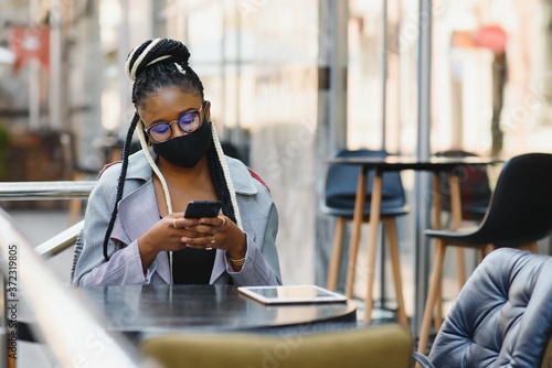 young pretty african american women in a medical protective mask outside in cafe. african girl with tablet and phone.