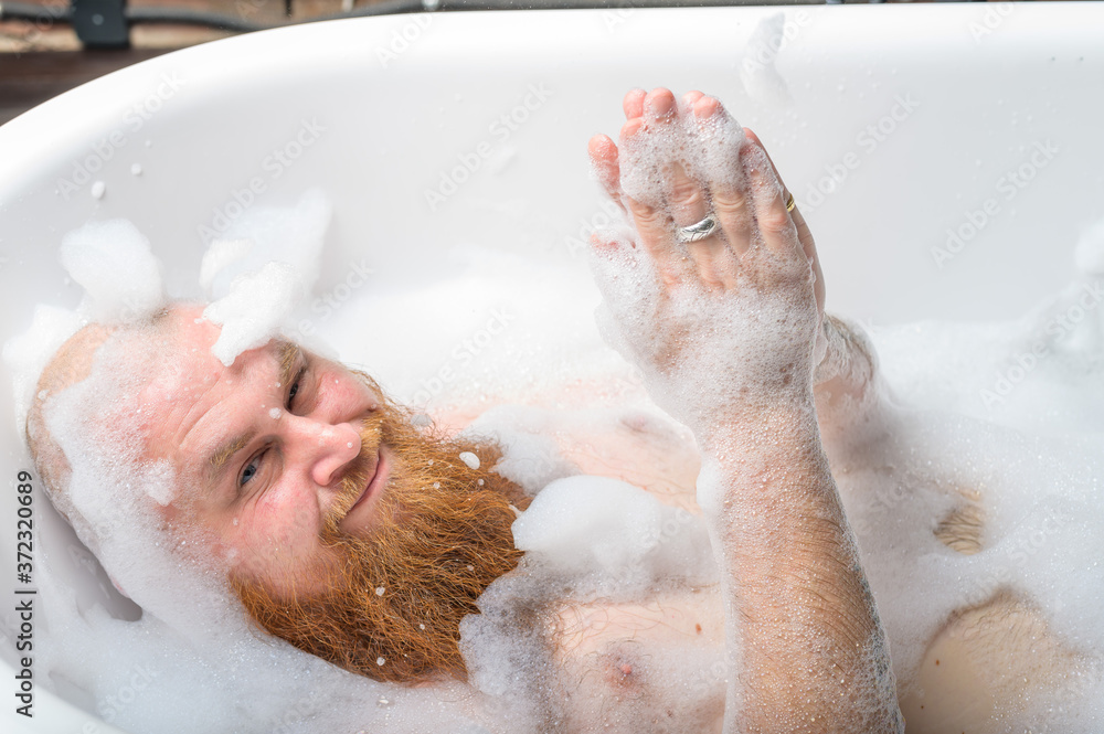 Close-up portrait of funny red-bearded bald man taking a bath with foam. Cheerful naughty guy at spa treatments. A parody of glamorous girls. Close-up portrait of funny red-bearded bald man taking a bath with foam. Cheerful naughty guy at spa treatments. A parody of glamorous girls.