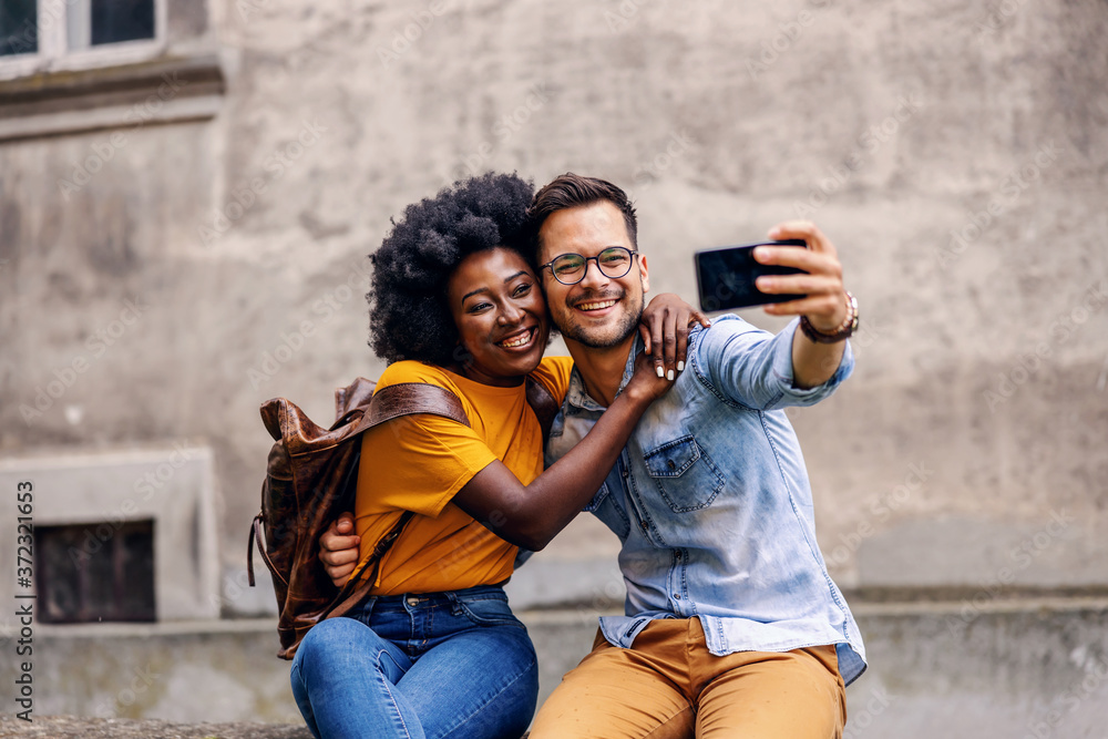 Young cute multicultural hipster couple hugging and taking selfie in an old part of the town.