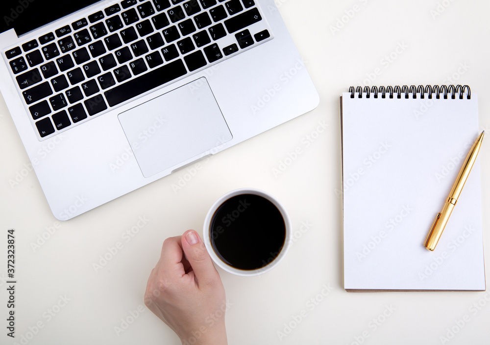 hand holds a cup of coffee on a working light table next to a laptop and accessories. notepad, golden pen. Top view with copy space for text input. hand holds a cup of coffee on a working light table next to a laptop and accessories. notepad, golden pen. Top view with copy space for text input.