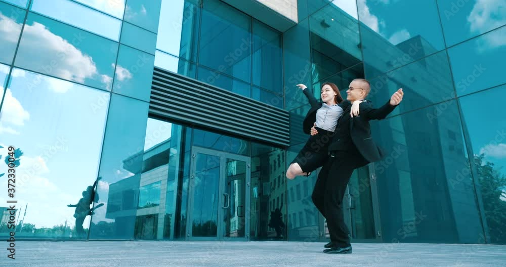 Couple of young successful office workers managers dancing on street near blue city building and flying papers. Students girl and guy celebrate victory, whirl hugging, man holds woman in his arms Couple of young successful office workers managers dancing on street near blue city building and flying papers. Students girl and guy celebrate victory, whirl hugging, man holds woman in his arms