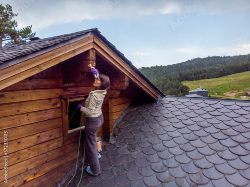 Woman on the roof of a chalet varnishing the wood safely tied with a rope