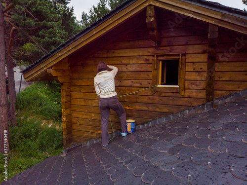 Woman standing up on a roof varnishing and painting the wall of a wooden chalet in the mountains