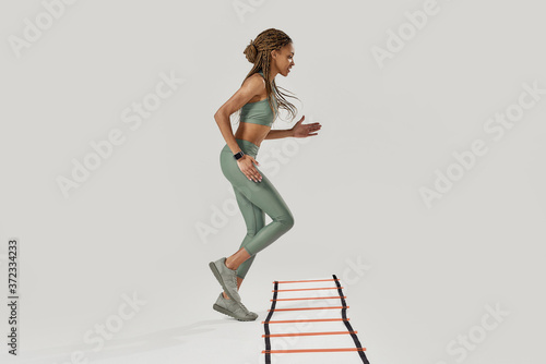 Fototapeta Naklejka Na Ścianę i Meble -  Life is movement. Full length shot of young sportive mixed race woman in sportswear training on agility ladder drill isolated over grey background. Fitness workout concept