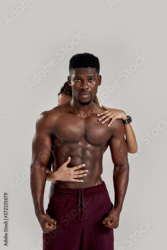 Muscular african american man looking at camera while sportive mixed race woman embracing, touching his body isolated over grey background. Sports, workout, bodybuilding concept