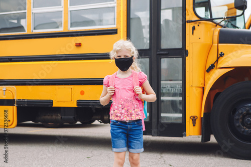 Happy Caucasian girl student wearing face mask near yellow bus. Kid with personal protective equipment on face. Education and back to school in September. A new normal during coronavirus.
