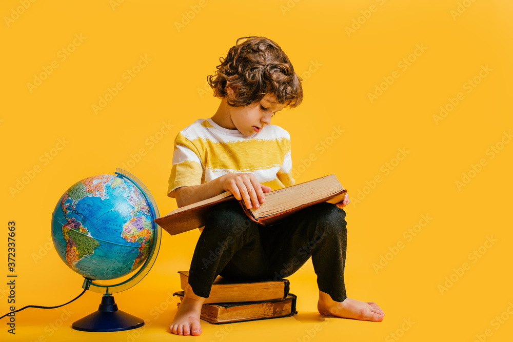 curly boy in glasses and stripes tshirt and sits on a stack of books, near to the globus on yellow backgroundm space for text. curly boy in glasses and stripes tshirt and sits on a stack of books, near to the globus on yellow backgroundm space for text.