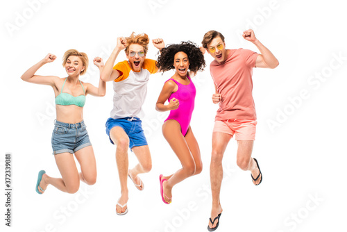 Multiethnic friends in sunglasses and swimsuits showing yeah gesture while jumping isolated on white