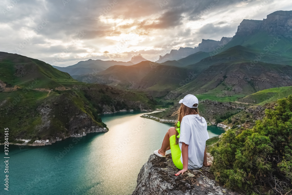 A beautiful girl tourist sits on a stone high in the mountains admiring the lake and sunset. The concept of tourism, travel and outdoor recreation