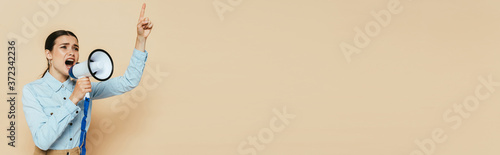 brunette woman in denim shirt screaming in loudspeaker on beige, panoramic shot