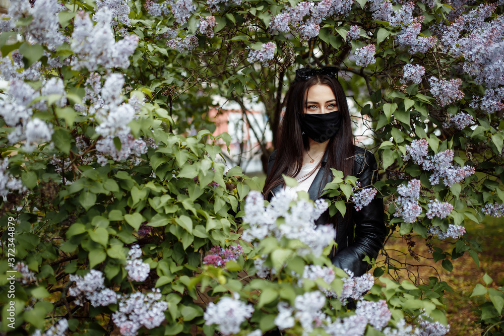Girl in a medical mask on the background of a blossoming lilac in the park. Black mask. Coronavirus protection. Spring allergy Girl in a medical mask on the background of a blossoming lilac in the park. Black mask. Coronavirus protection. Spring allergy