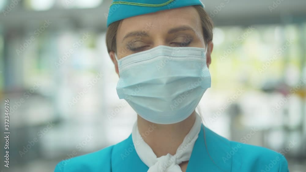 Close-up portrait of serious Caucasian stewardess in face mask looking at camera. Professional female flight attendant posing in airport during coronavirus pandemic. Covid-19 international tourism. Close-up portrait of serious Caucasian stewardess in face mask looking at camera. Professional female flight attendant posing in airport during coronavirus pandemic. Covid-19 international tourism.