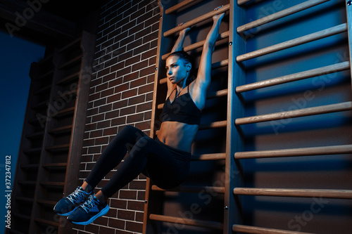 Confident fit young brunette woman doind exercises for abs on the wall bars in gym in neon lights.