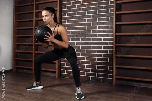 Fit young brunette woman doing squat exercise with medicine ball in gym.
