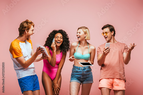 excited africal american woman talking on smartphone near multicultural friends using gadgets isolated on pink photo