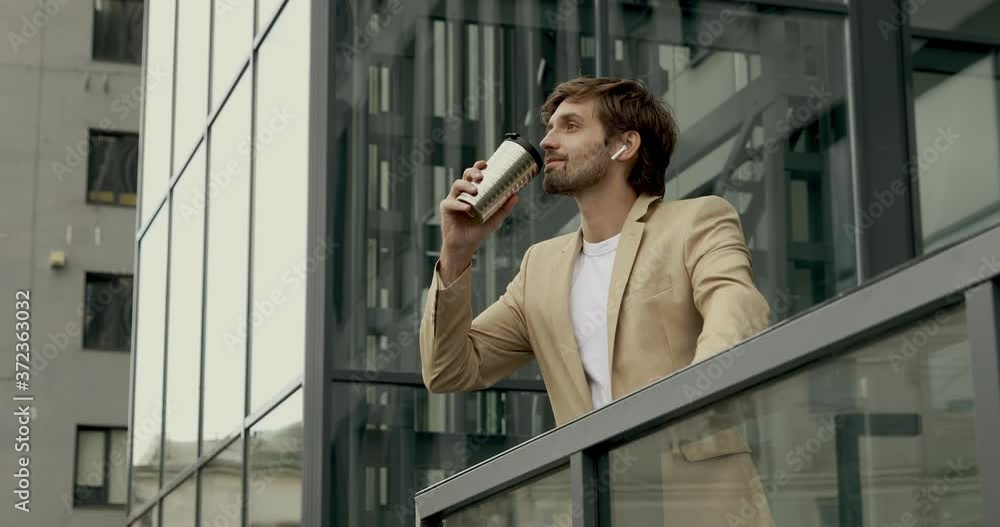 Happy bearded man in stylish outfit standing near office center and drinking hot beverage from thermo cup. Young businessman enjoying coffee break on fresh air. Happy bearded man in stylish outfit standing near office center and drinking hot beverage from thermo cup. Young businessman enjoying coffee break on fresh air.