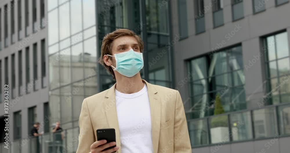 Handsome man in formal clothing and medical mask using modern smartphone while walking on street. Concept of pandemic, work and technology. Handsome man in formal clothing and medical mask using modern smartphone while walking on street. Concept of pandemic, work and technology.
