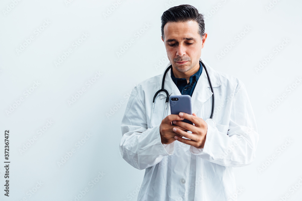 Black haired doctor wearing white coat and stethoscope looking at his mobile phone, on white background. Medicine concept Black haired doctor wearing white coat and stethoscope looking at his mobile phone, on white background. Medicine concept