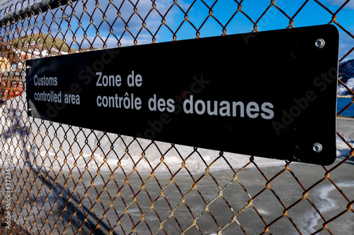 A black and white fence with a sign attached to a chain link industrial boarder. It has customs controlled area in white text. There's a harbour with a wharf in the background under a blue sky.