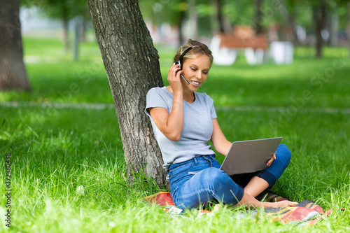 Smiling woman with laptop and headset having online conversation while sitting in city park. © Stavros