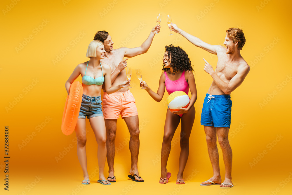 Obraz premium full length view of shirtless man clinking champagne glasses near multicultural women with inflatable ball and ring on yellow