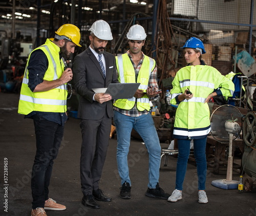 Group of factory workers consists of Manager, Engineers, Technicians with hardhat and vest jacket, discussing about industrial factory production management by using computer notebook for information