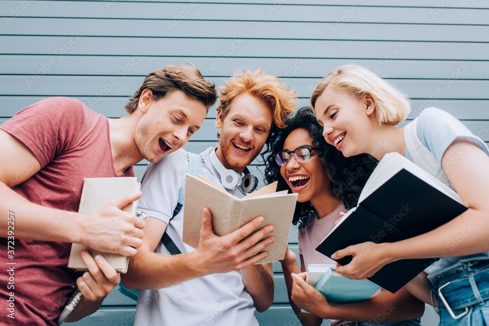 Obraz premium low angle view of excited multicultural students reading book on urban street