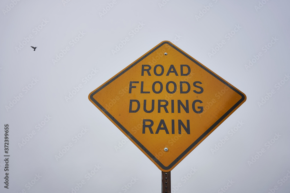 'Road Floods During Rain' warning sign against overcast sky in the rain.