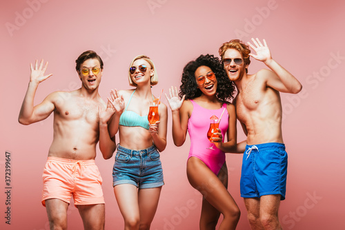 young multicultural friends in summer outfit waving hands at camera while holding cocktail glasses isolated on pink