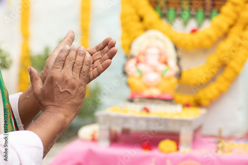 Close up of Elder man hands offering Bhajan or hymn in front of Lord Ganesha Idol by clapping during Ganesha or vinayaka Chaturthi festvial ceremony at home.