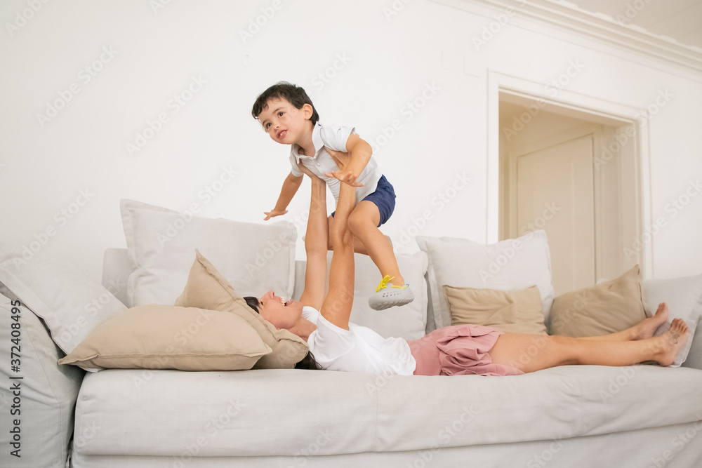 Happy young mother lying on sofa and playing with son. Beautiful Caucasian mom holding cute little boy with hands and laughing in living room. Motherhood, love, family time and leisure concept Happy young mother lying on sofa and playing with son. Beautiful Caucasian mom holding cute little boy with hands and laughing in living room. Motherhood, love, family time and leisure concept