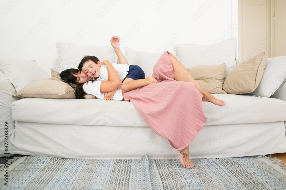 Caucasian mother laying on couch, hugging little son and talking with kid. Long-haired pretty mom with tattoo on foot. Cute boy sitting on woman chest. Family, love and togetherness concept Caucasian mother laying on couch, hugging little son and talking with kid. Long-haired pretty mom with tattoo on foot. Cute boy sitting on woman chest. Family, love and togetherness concept