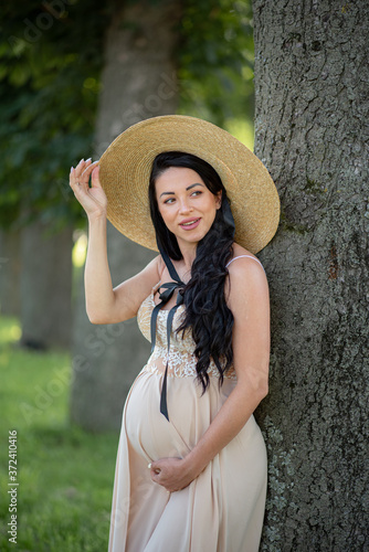 Pregnant woman posing in a beige dress on a background of green trees.