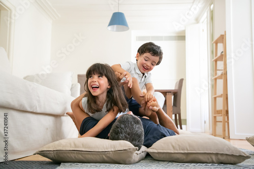 Fotografía Happy dad and excited little kids having fun together, romping and playing on living room carpet, enjoying time at home
