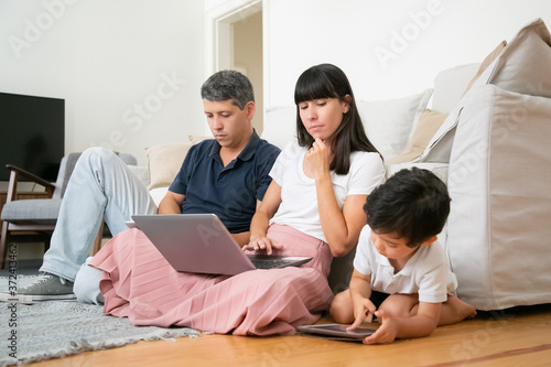 Mom with laptop watching little son using tablet, sitting on floor in living room by his parents. Digital gadgets and family concept © Mangostar