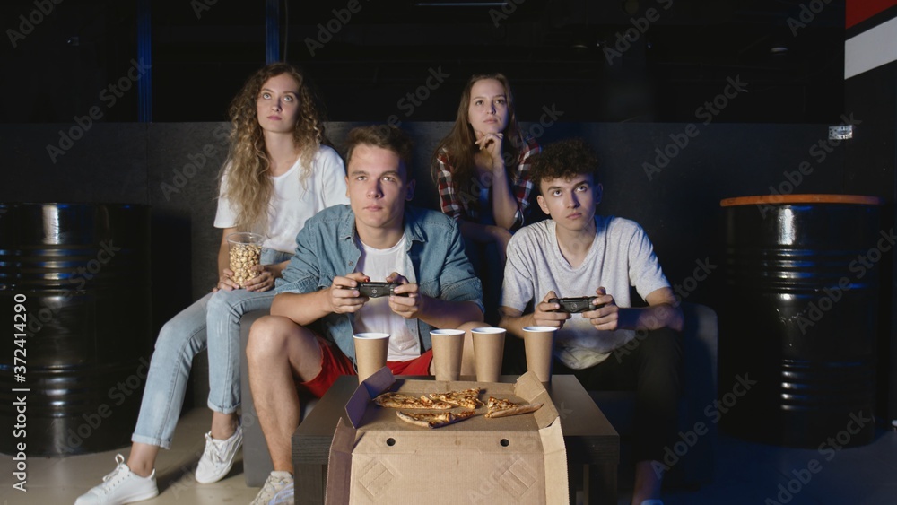 Young friends playing on the game console. Two men holds controllers and plays while girlfriends watches Young friends playing on the game console. Two men holds controllers and plays while girlfriends watches