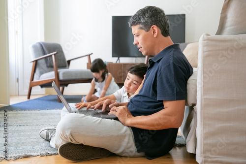 Focused dad and little son sitting on floor in apartment, using laptop, working or watching content. Communication or technology concept