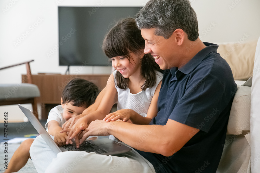 Fototapeta premium Joyful happy dad and two kids using laptop together, sitting on floor in apartment, pressing buttons in keyboards. Leisure time with family and technology concept