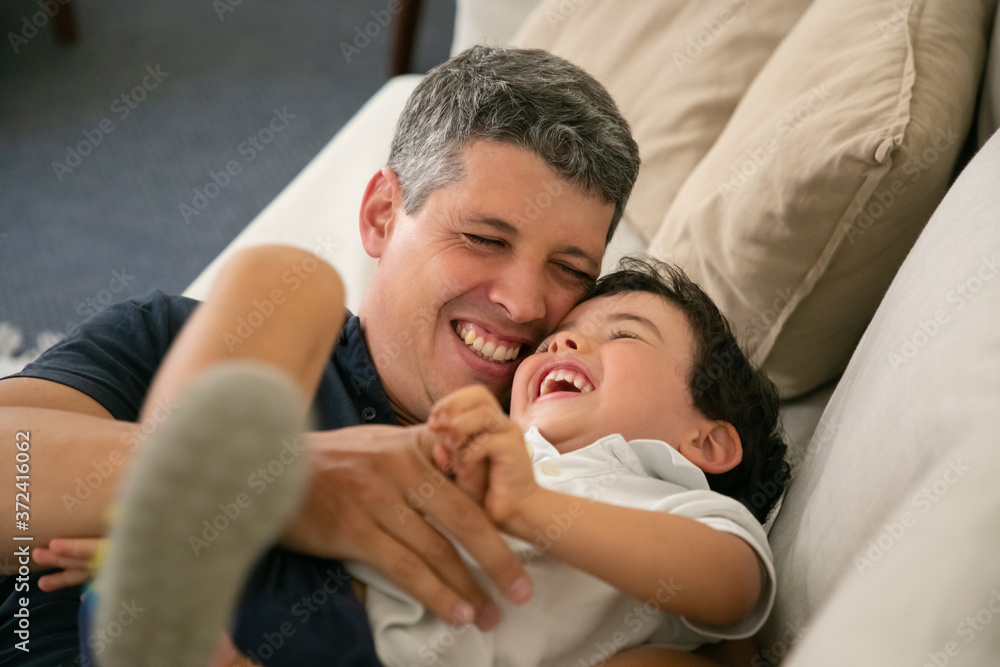 Obraz premium Laughing father tickling his adorable little boy on sofa. Happy attractive Caucasian dad hugging or embracing his lovely son, smiling and playing with child. Fatherhood, leisure and parenthood concept